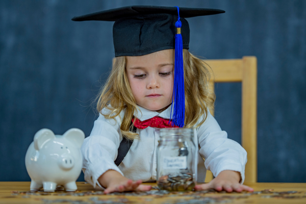 Little girl with a graduation cap on sitting at a table with a piggy bank on it and lots of change in front of her.